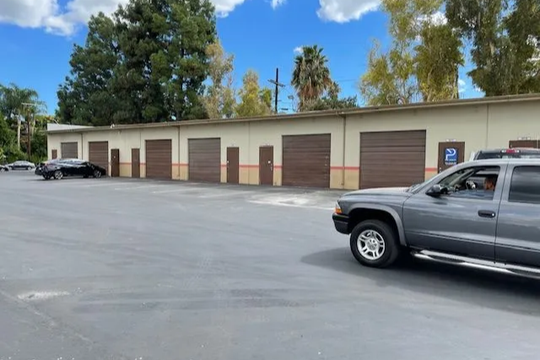 A row of storage units with brown doors sits in front of an open parking area, with two cars parked nearby under a blue sky with scattered clouds.