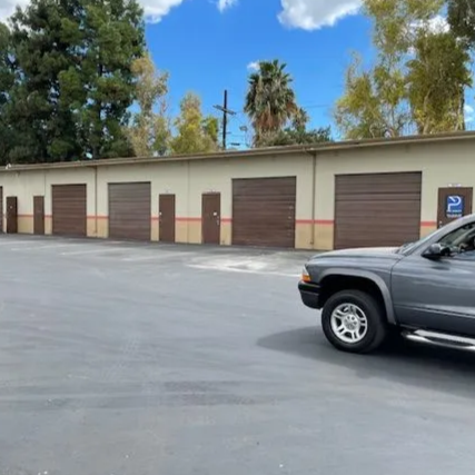 A row of storage units with brown doors sits in front of an open parking area, with two cars parked nearby under a blue sky with scattered clouds.