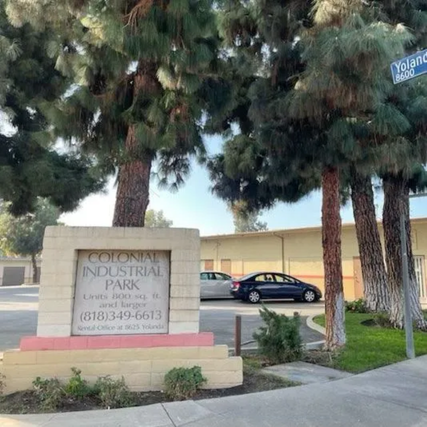 A photo of tall pine trees standing beside a street corner with a sign that reads “Colonial Industrial Park” in front of a low industrial building, with a parked car visible in the background.
