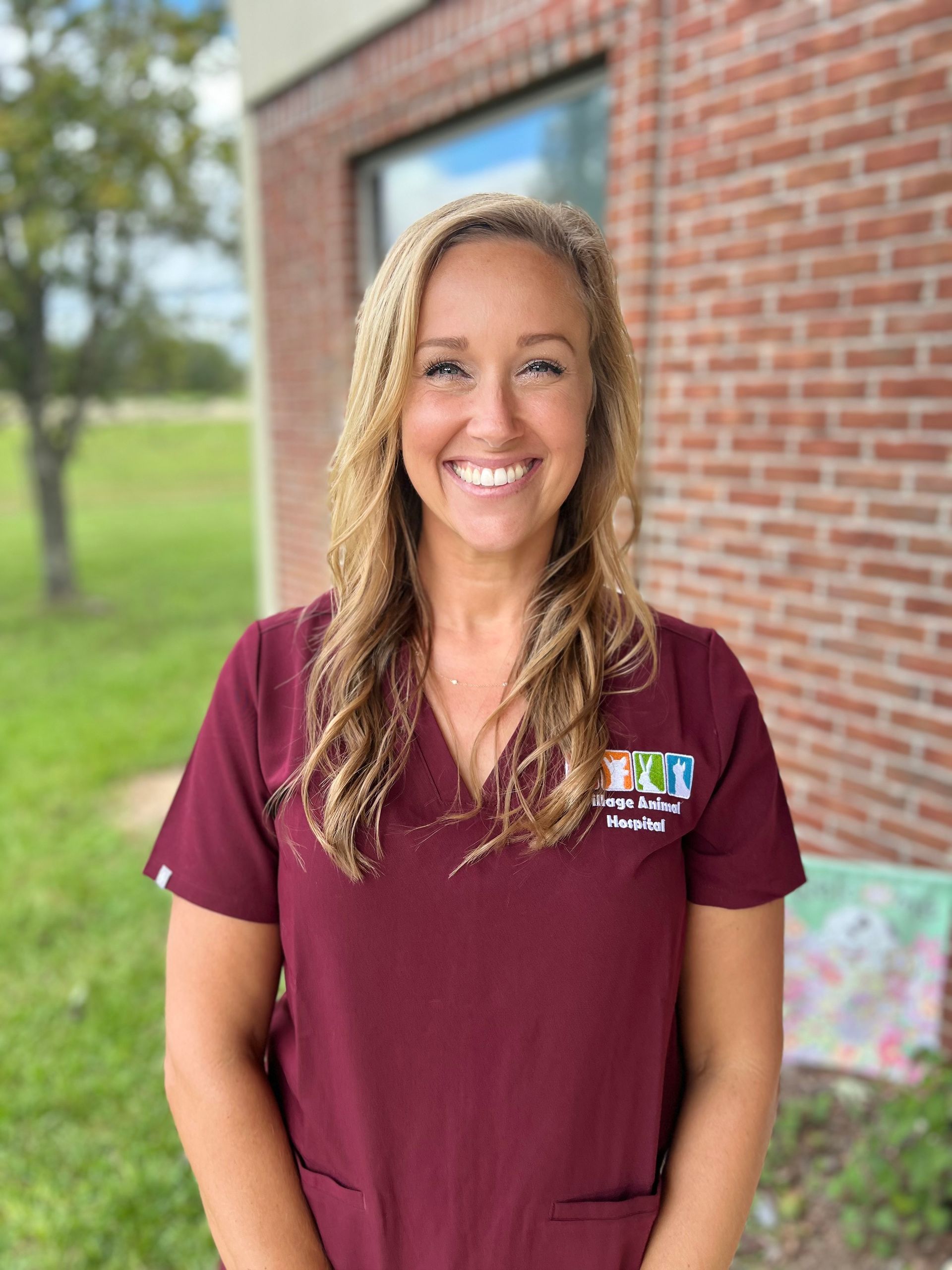 A woman in a maroon scrub top is smiling in front of a brick building.