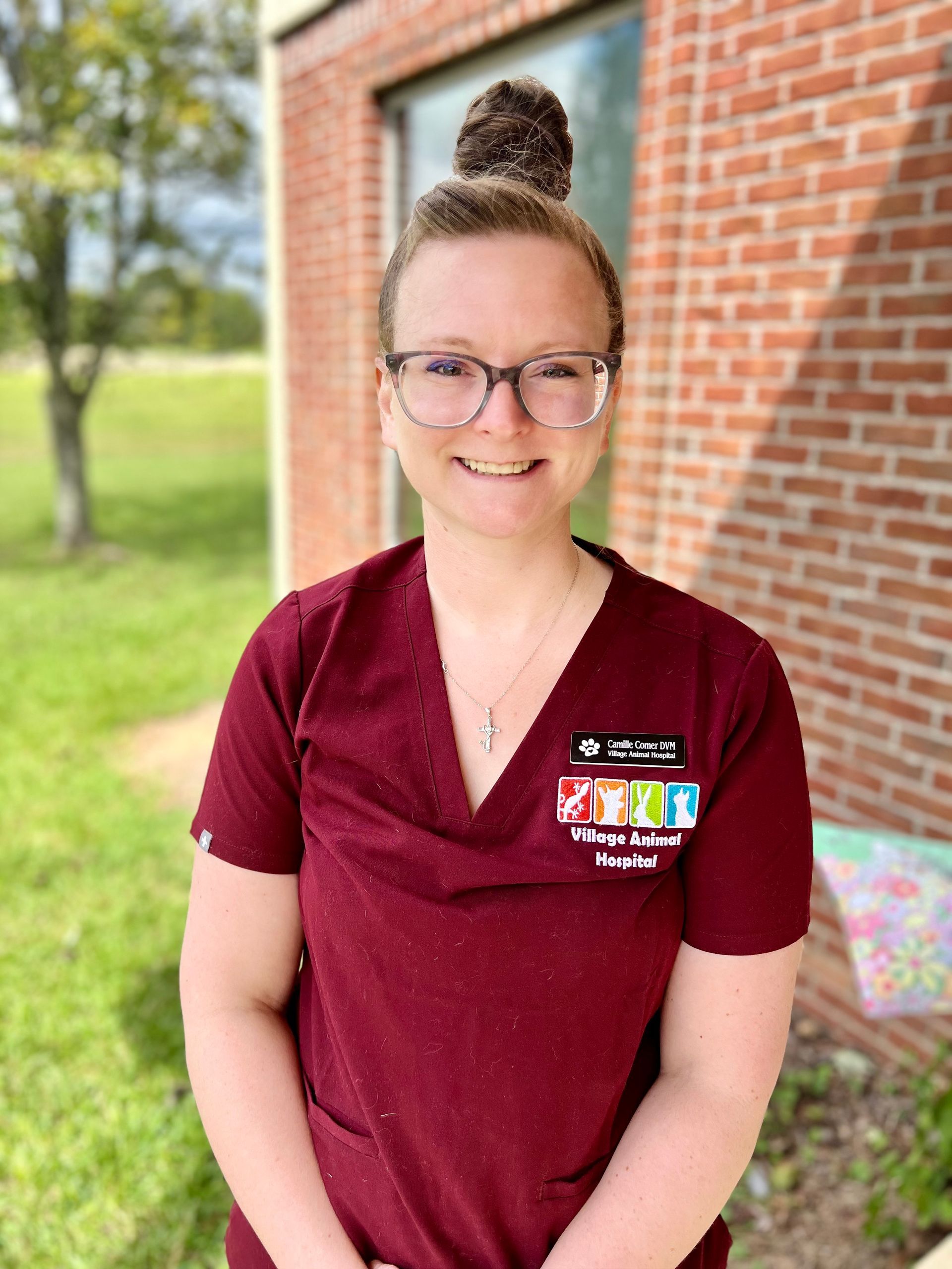 A woman wearing glasses and a maroon scrub top is standing in front of a brick building.