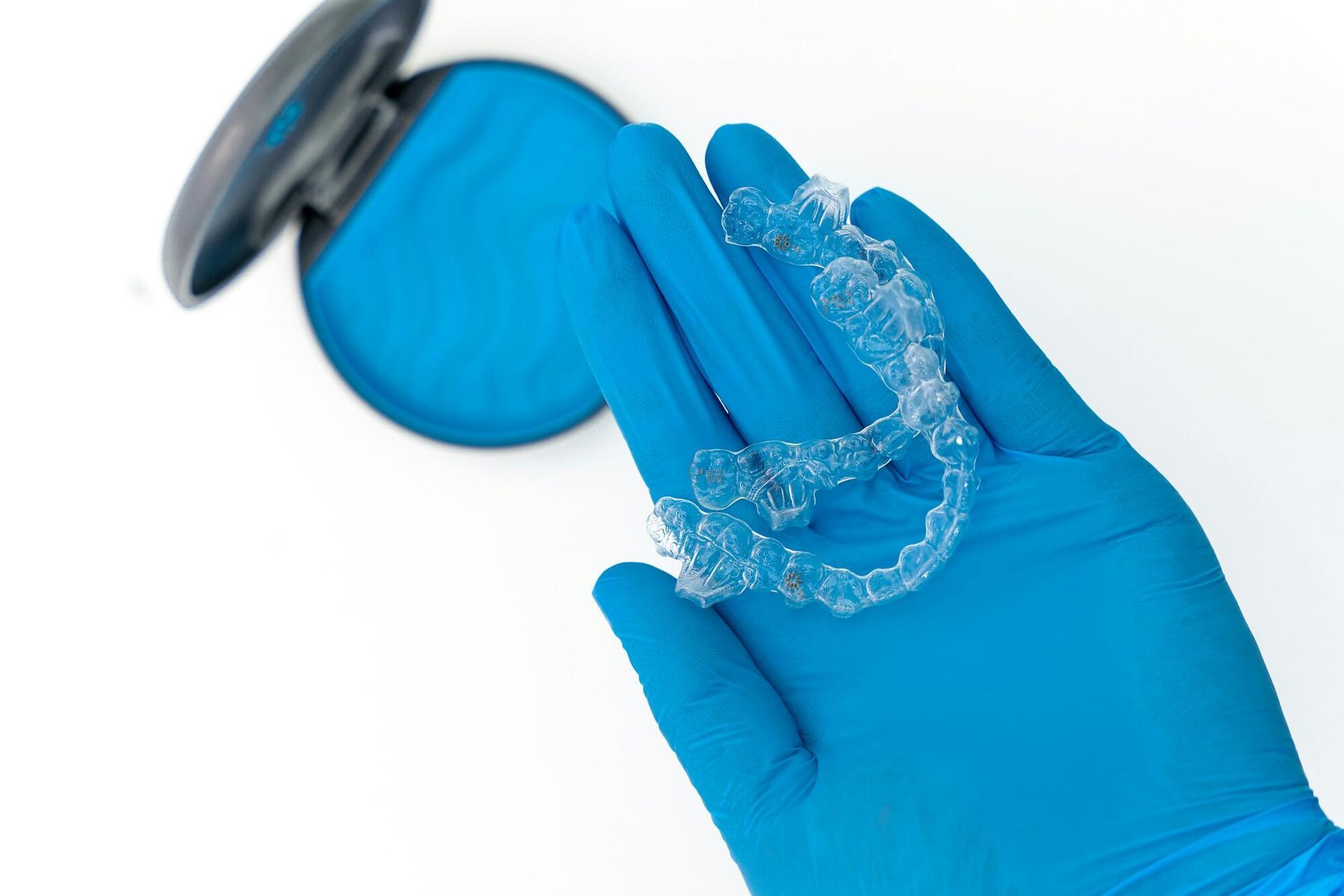 Gloved hand holding clear aligners, next to an open blue case, against a white background.