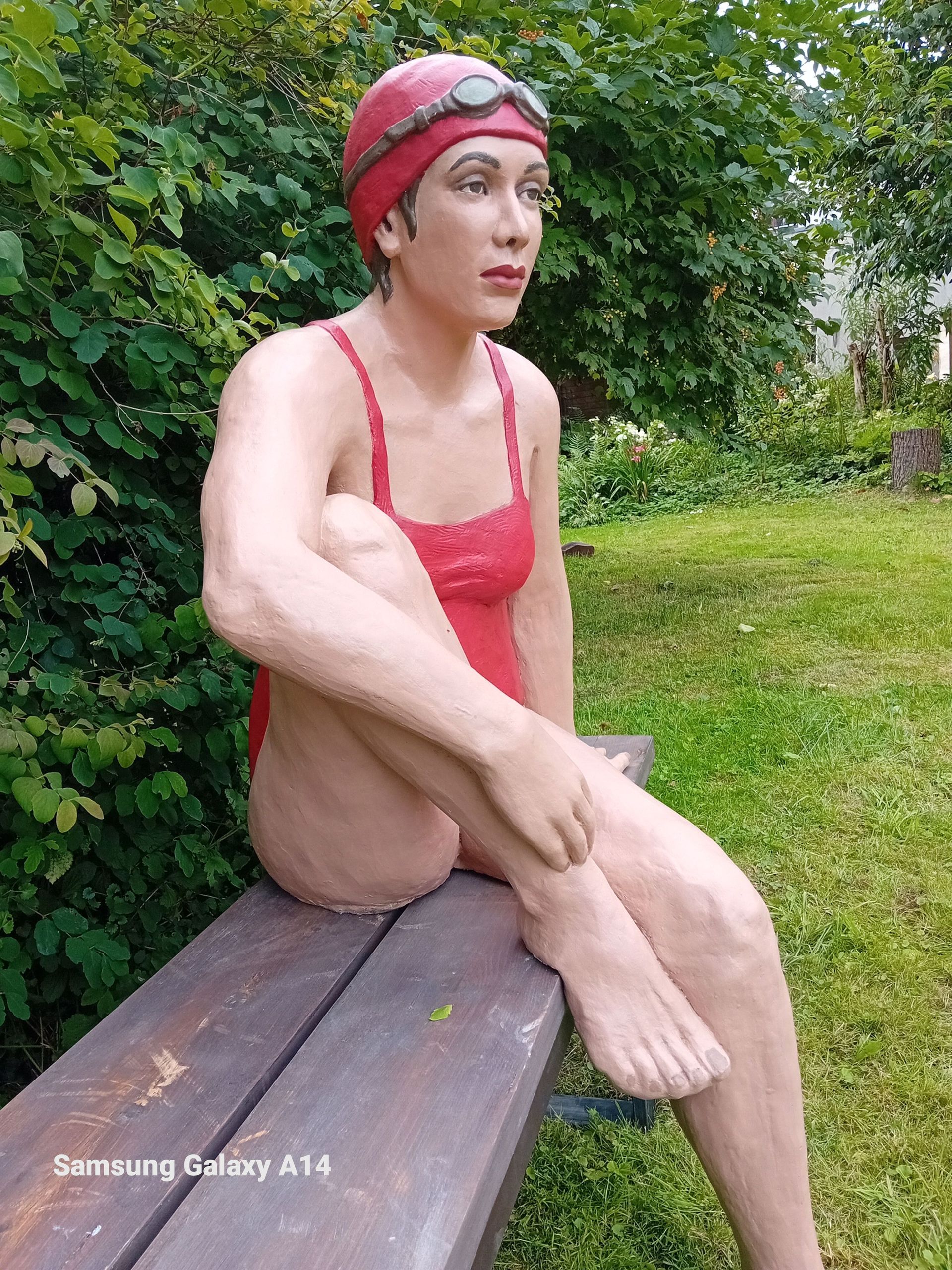 Eine Statue einer Frau in einem roten Badeanzug sitzt auf einer Holzbank.