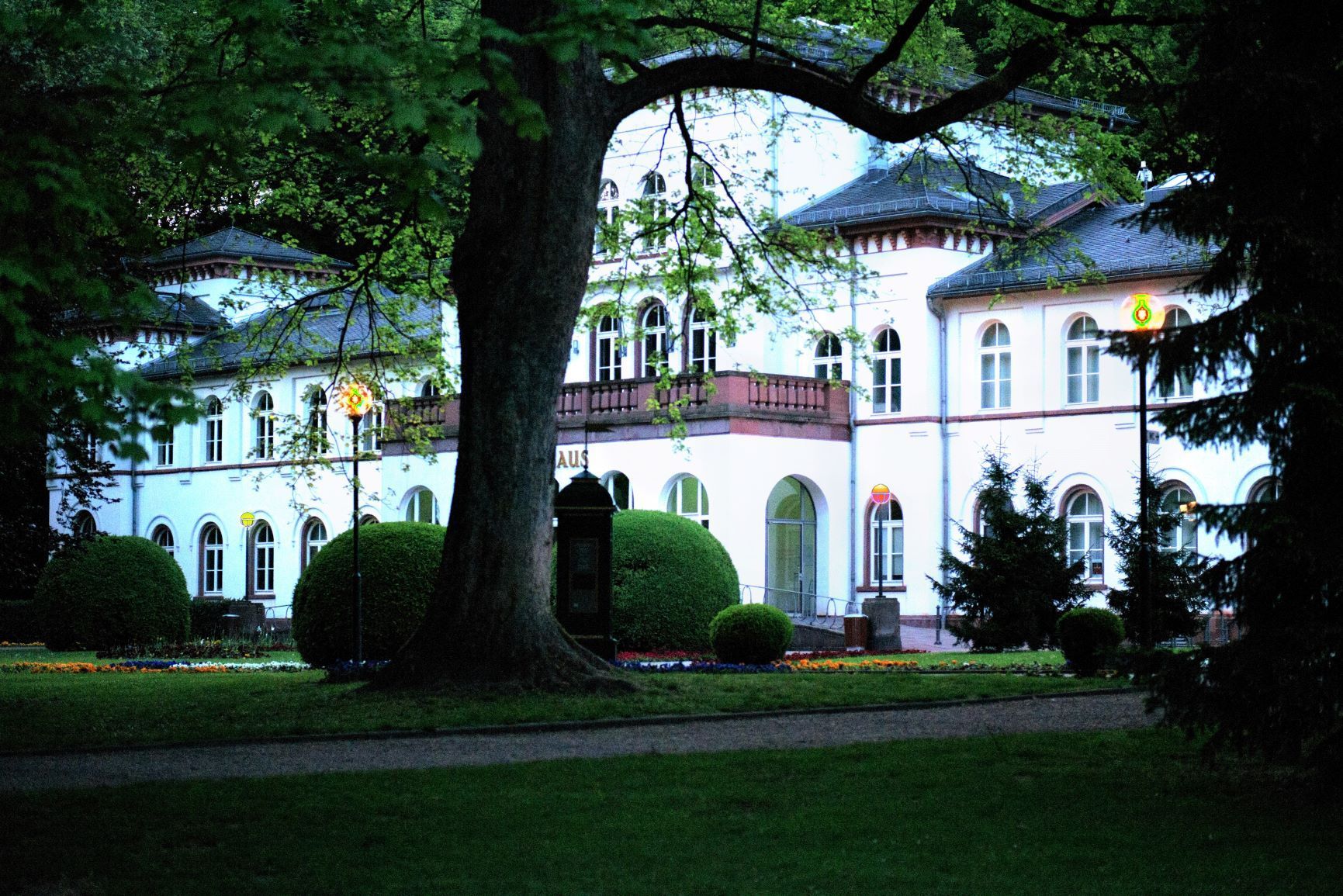 Weißes Gebäude im europäischen Stil mit mehreren Bogenfenstern und Rundtürmen, teilweise von Bäumen verdeckt; grüner Rasen.