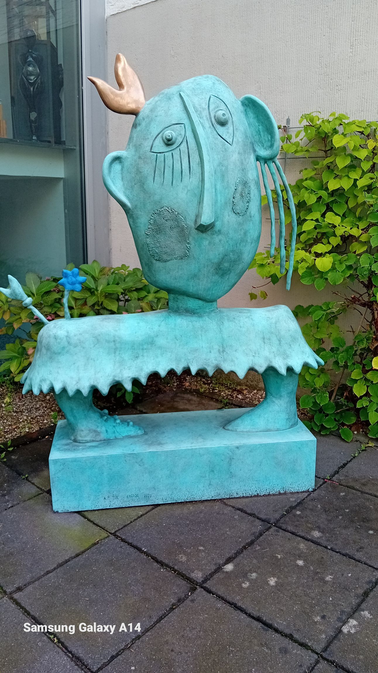 Blaugrüne Skulptur einer Person mit Krone und langen Ohren auf einem gefliesten Gehweg.