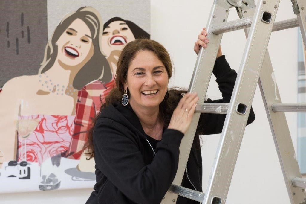 Eine Frau lächelt neben einer Leiter und einem Gemälde mit lachenden Menschen.