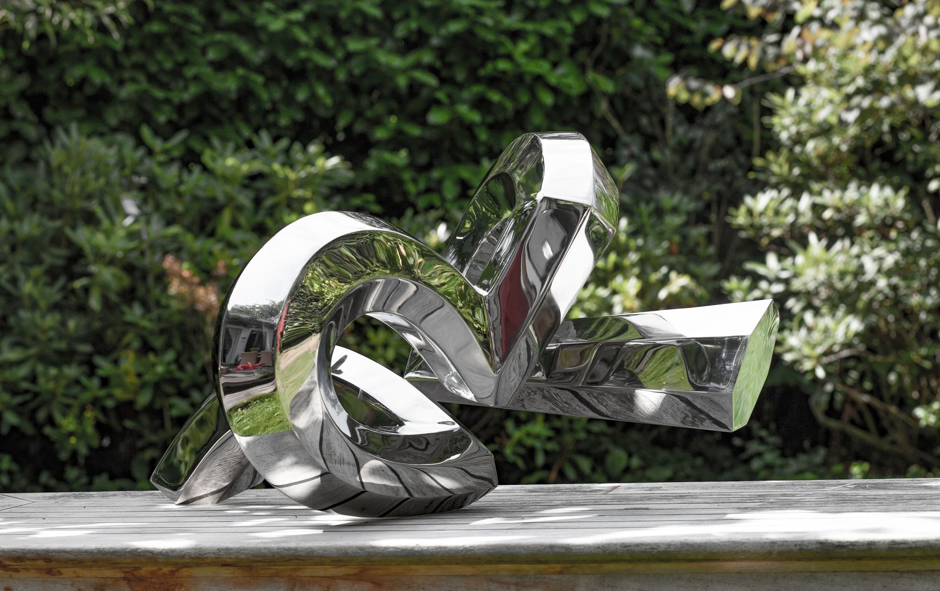 Eine glänzende, geknotete Metallskulptur im Freien, die die Umgebung reflektiert, vor einem Hintergrund aus grünem Laub.