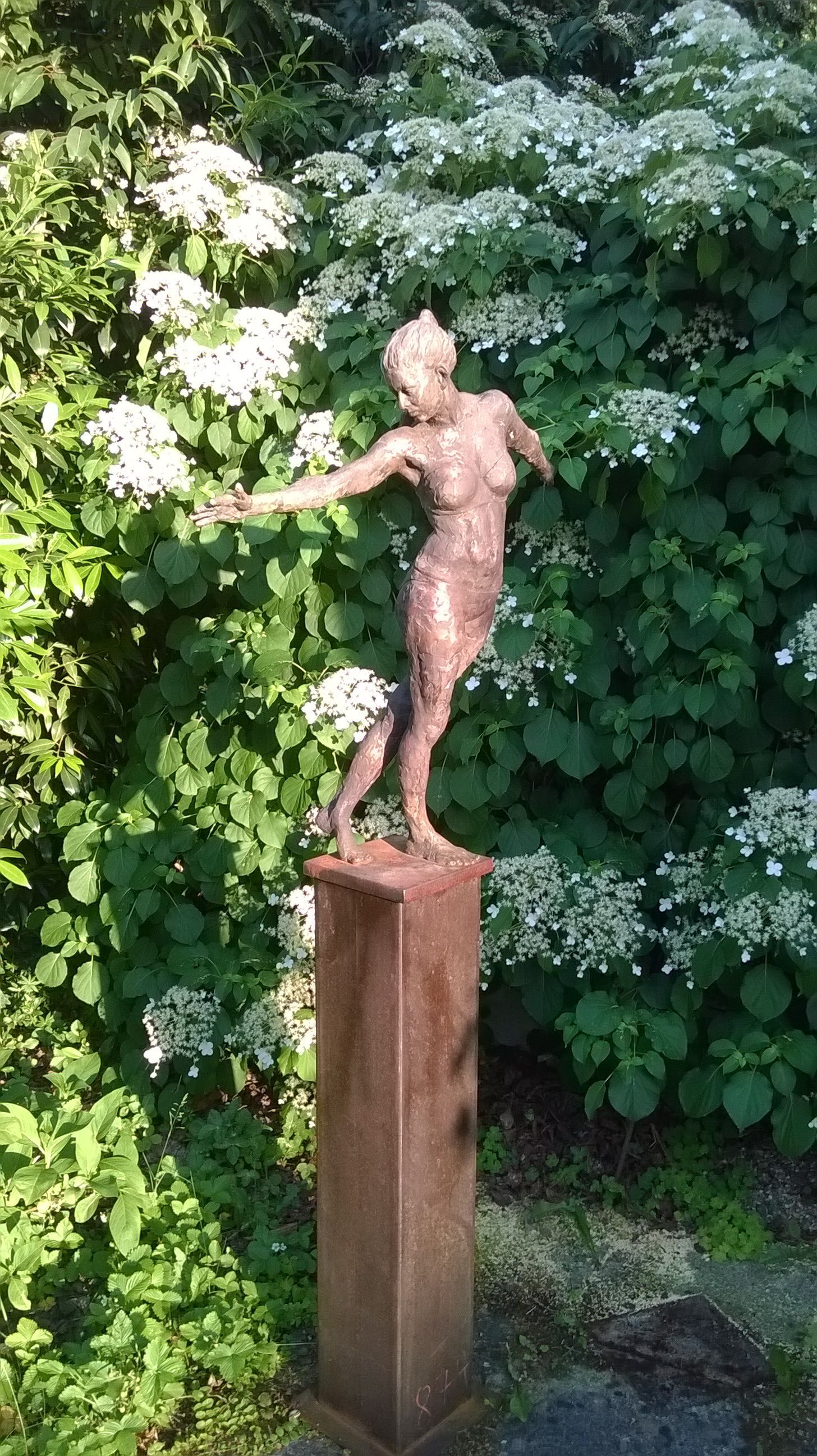 Bronzestatue einer nach oben ausgestreckten Frau, auf einer quadratischen Säule, vor weißen Blüten und grünem Laub.