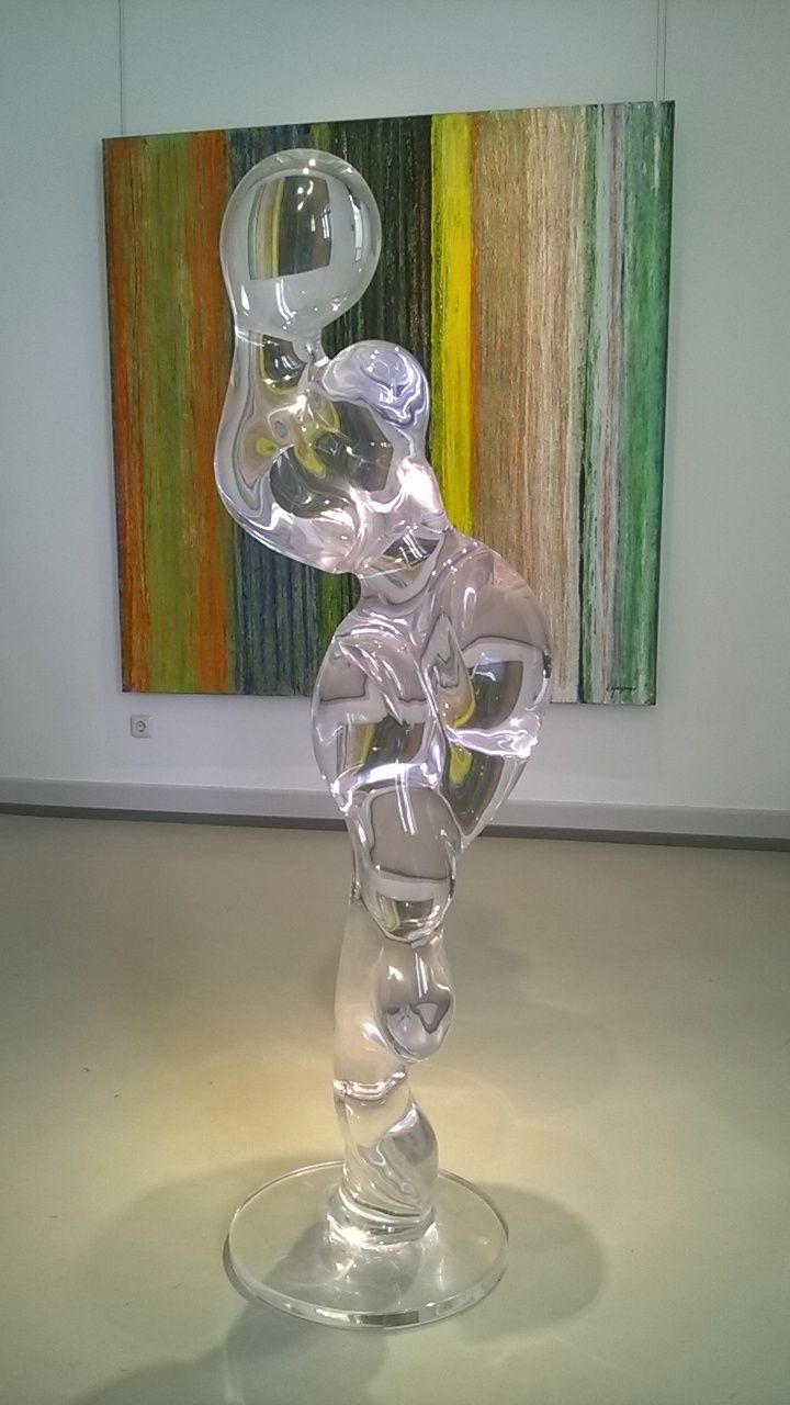 Klare Glasskulptur einer Figur mit geschwungenen Formen auf einem Sockel, vor einem farbenfrohen, gewebten Hintergrund.