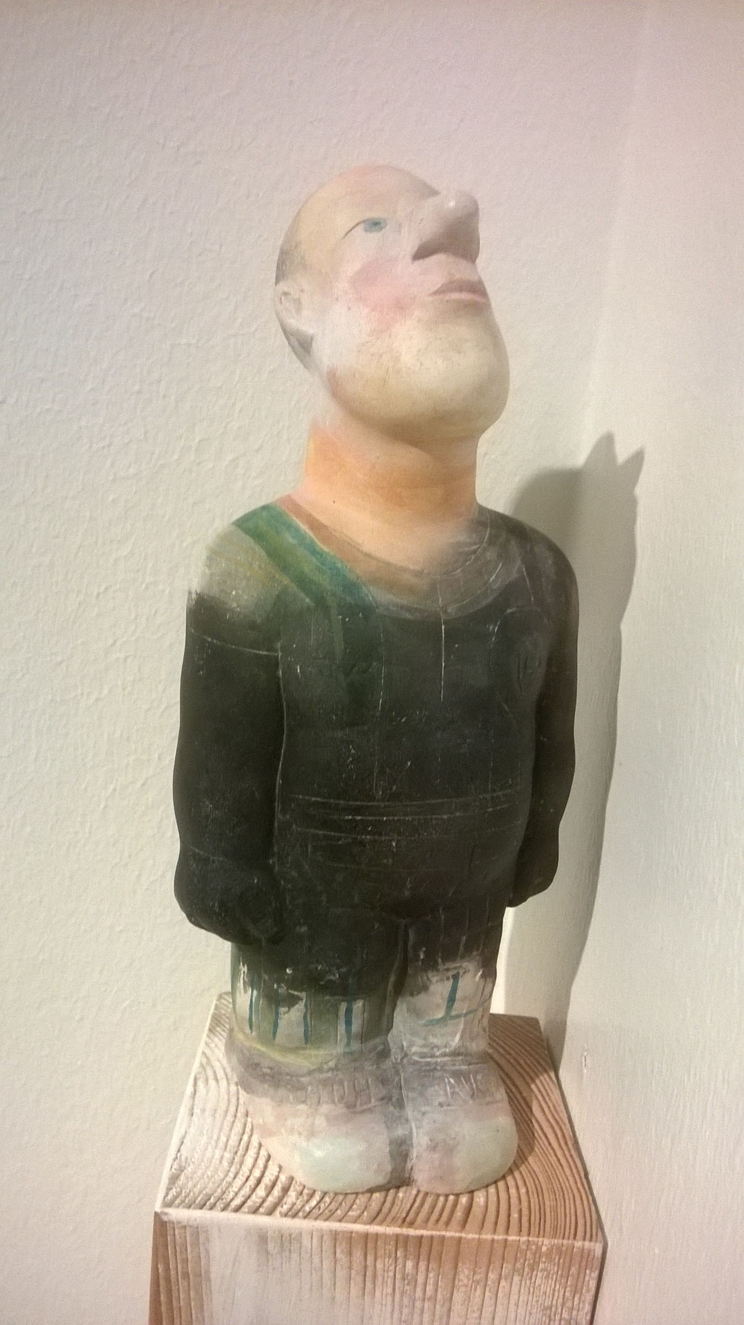 Skulptur eines Mannes, der nach oben blickt, ein grünes Hemd, einen Bart und weiße Stiefel trägt, auf einem Holzsockel.