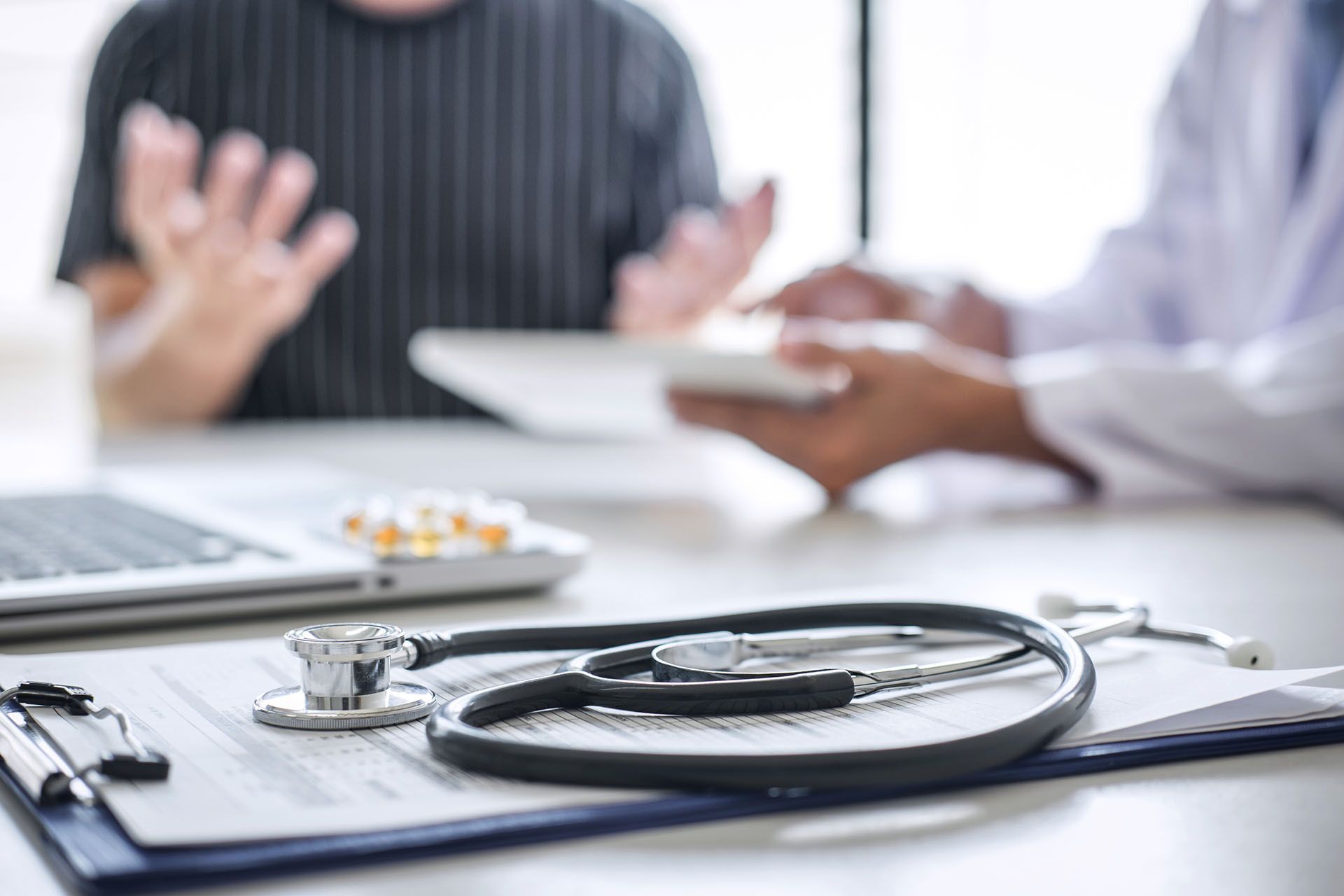 Stethoscope, clipboard, and pills on desk with doctor and patient blurred in background.