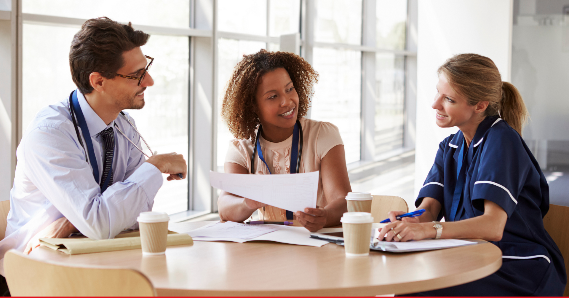 Medical professionals in a meeting at a table, discussing papers, near a window.
