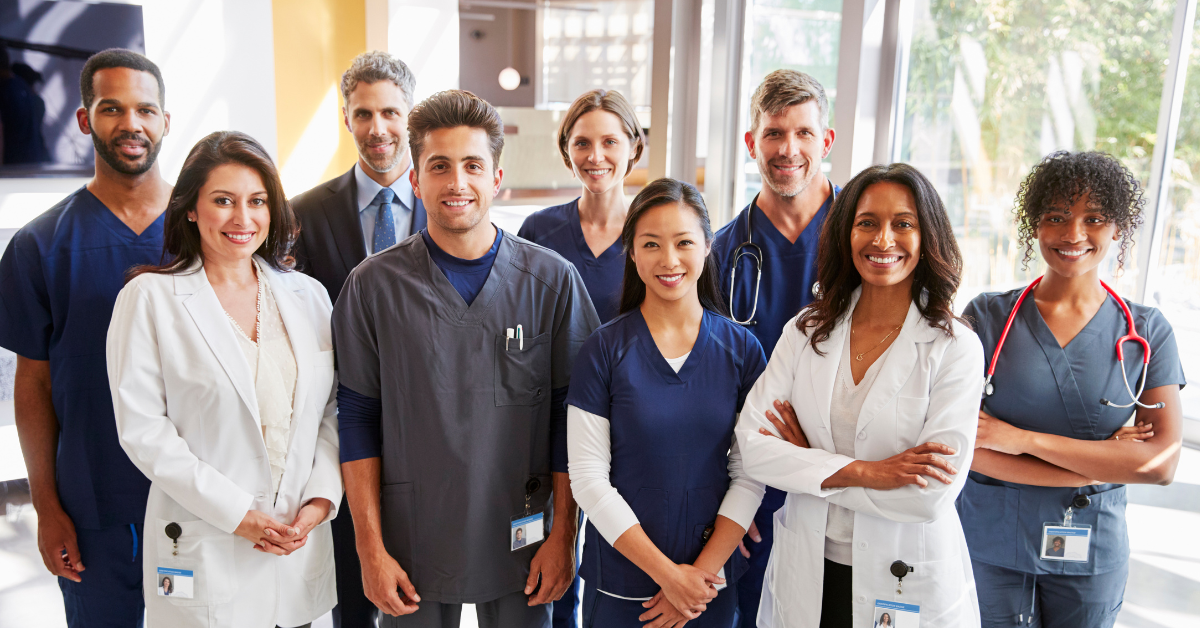 Medical professionals in scrubs and white coats stand together smiling in a modern hallway.