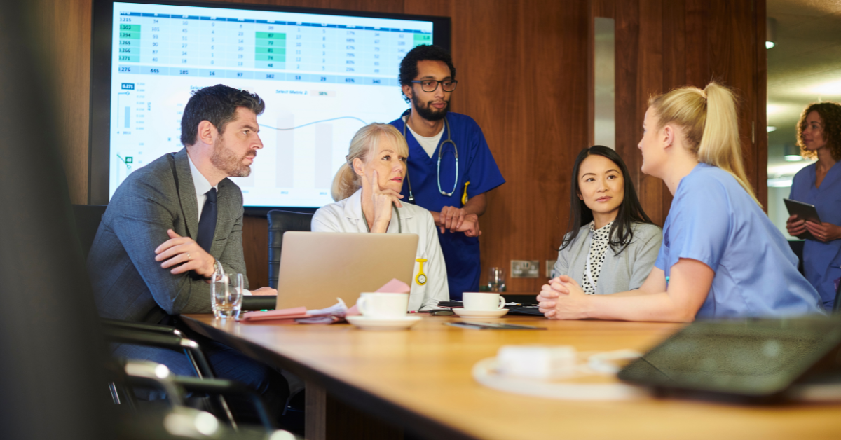 Group of people at a conference table with a screen showing data, discussing healthcare information.
