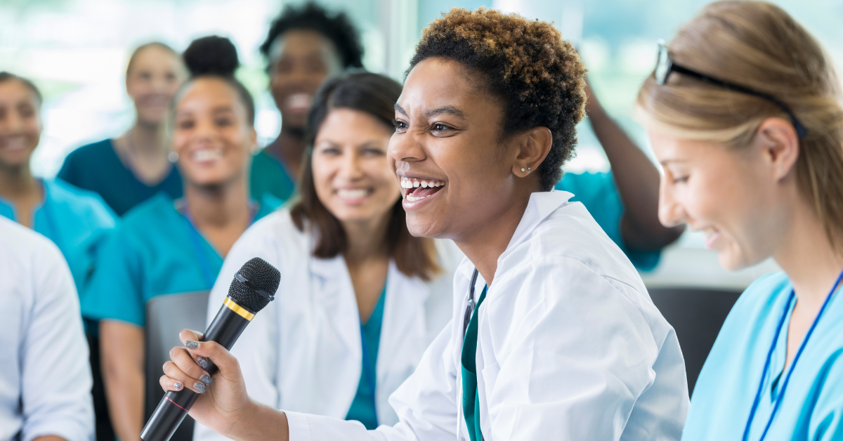 Woman in lab coat speaking into a microphone, surrounded by smiling medical professionals.