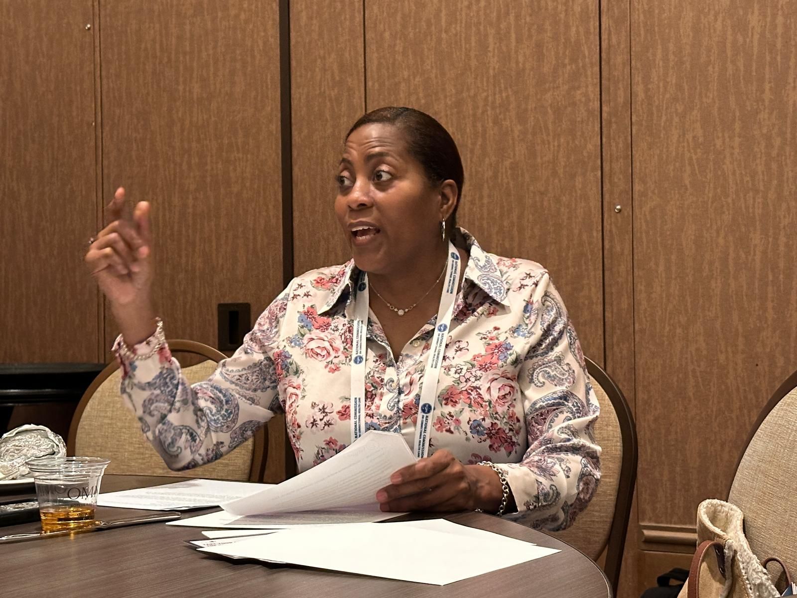 Woman seated at a table, gesturing and speaking, holding papers. Light-colored floral shirt. Interior setting.