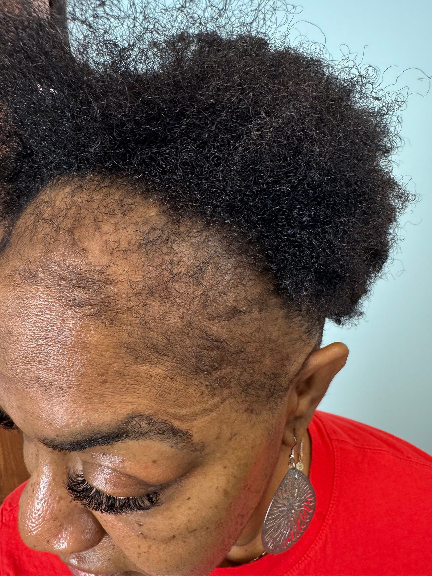 Close-up of a person with short, dark, textured hair; the scalp is visible. The person wears a red shirt and earrings.