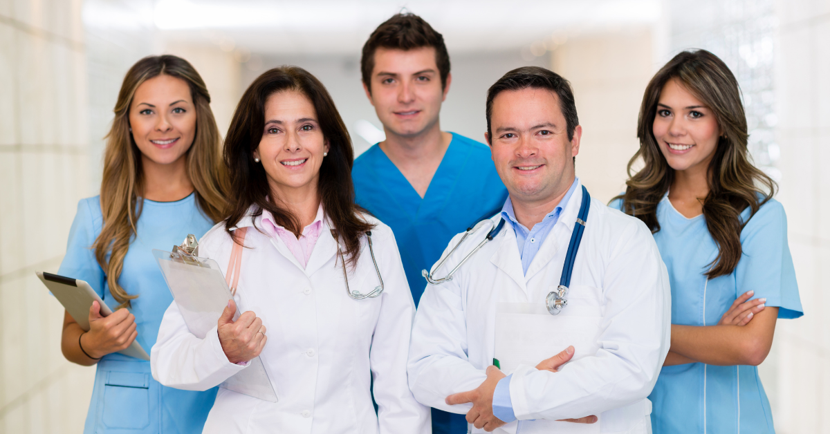 Medical team smiling in a brightly lit hallway. Three wear white coats, two wear scrubs; all have stethoscopes.