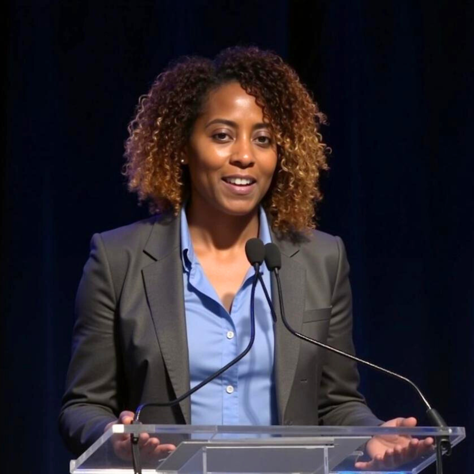 Woman speaking at a podium, wearing a blue shirt and grey blazer, in front of a dark backdrop.