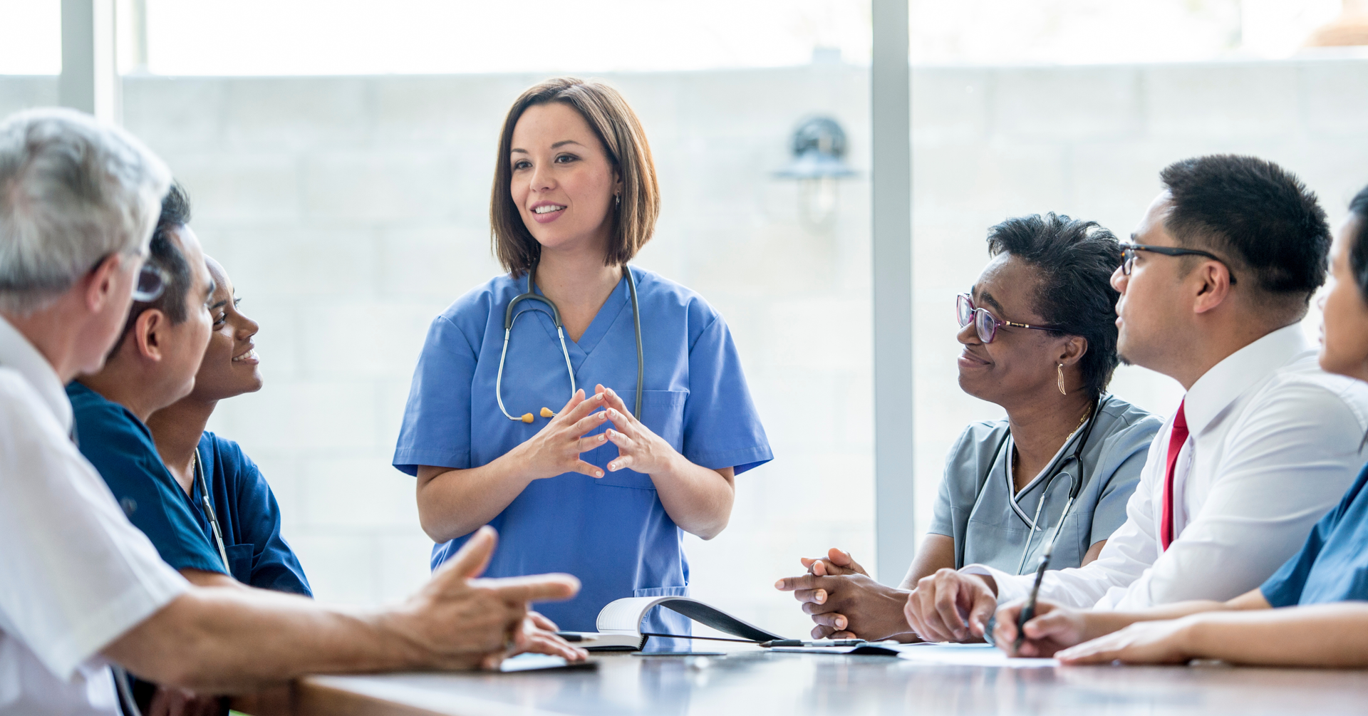 Medical professionals in scrubs and business attire in a meeting, with a woman in scrubs speaking.