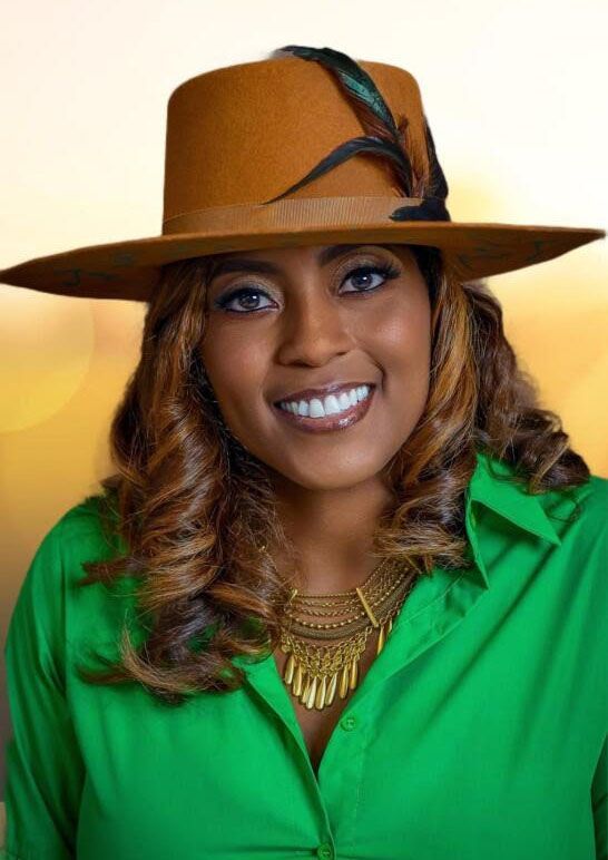 Woman in brown hat with feather, green shirt, gold necklace smiles. Soft, sunny background.