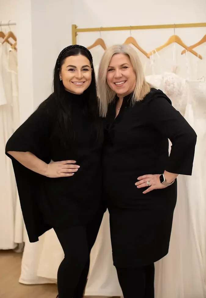 Two women in black clothing smiling in a bridal shop, dresses hanging in the background.