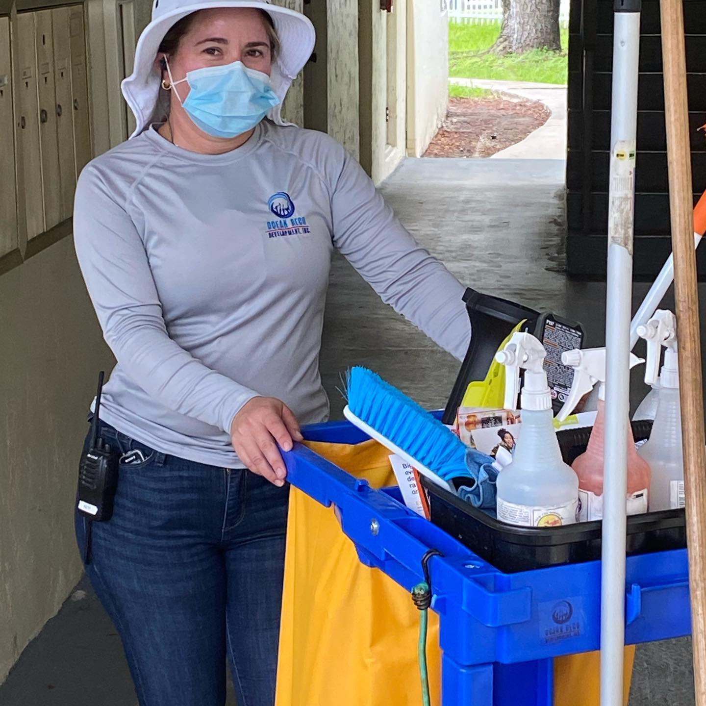 A woman wearing a mask and a hat is standing next to a cart full of cleaning supplies.