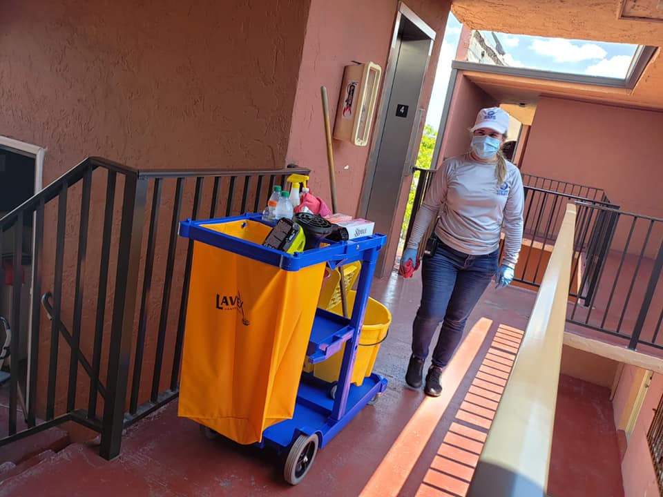 A woman wearing a mask is pushing a cleaning cart down a hallway.