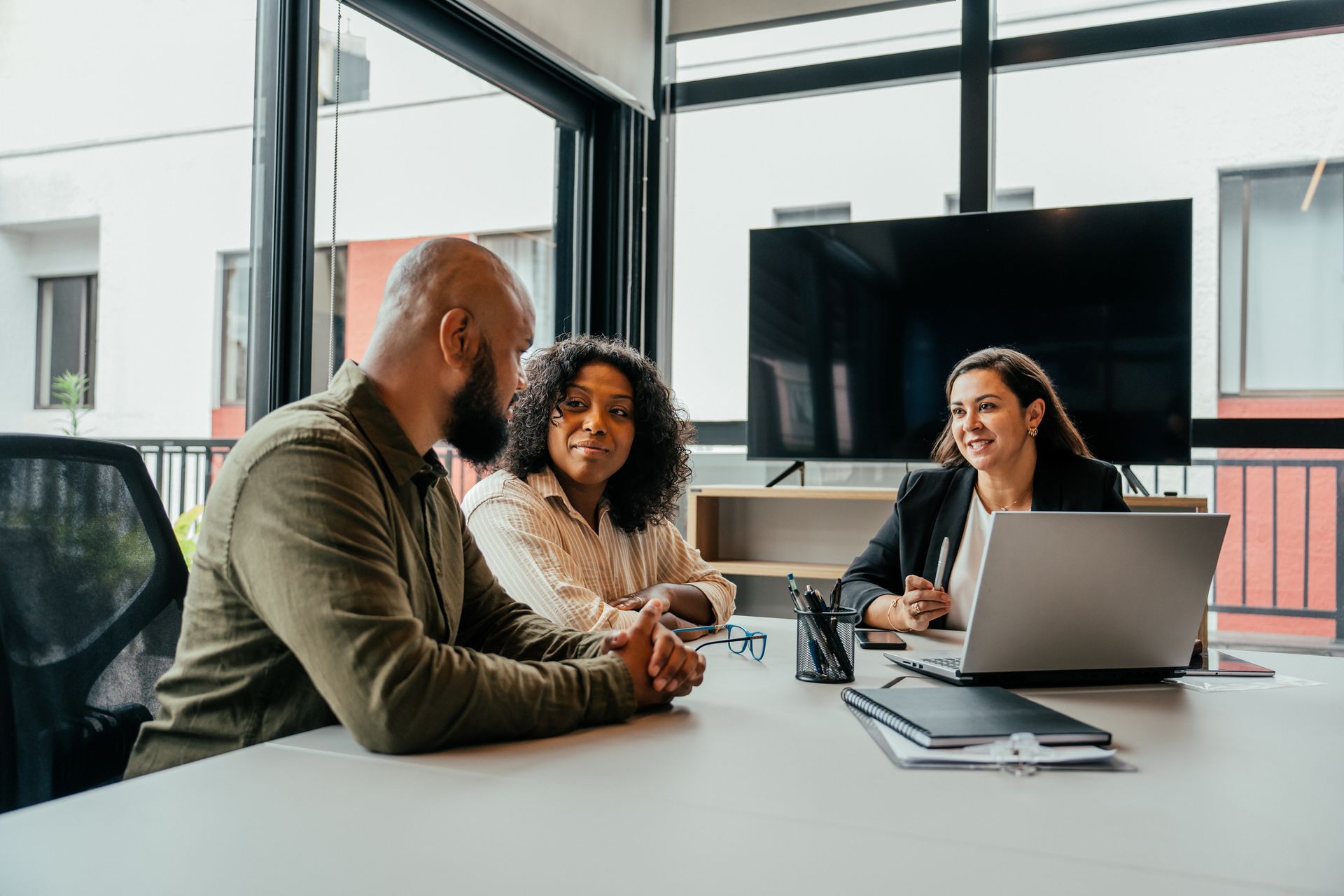 Three people in a business meeting: a man and woman listening to a woman at a laptop, indoors at a table.