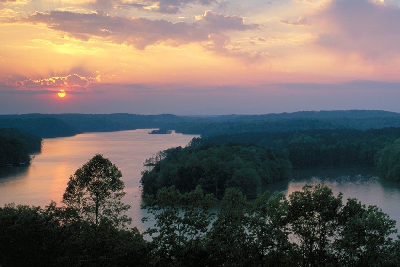 Sunset over a lake surrounded by trees; orange and pink sky reflecting in the water.