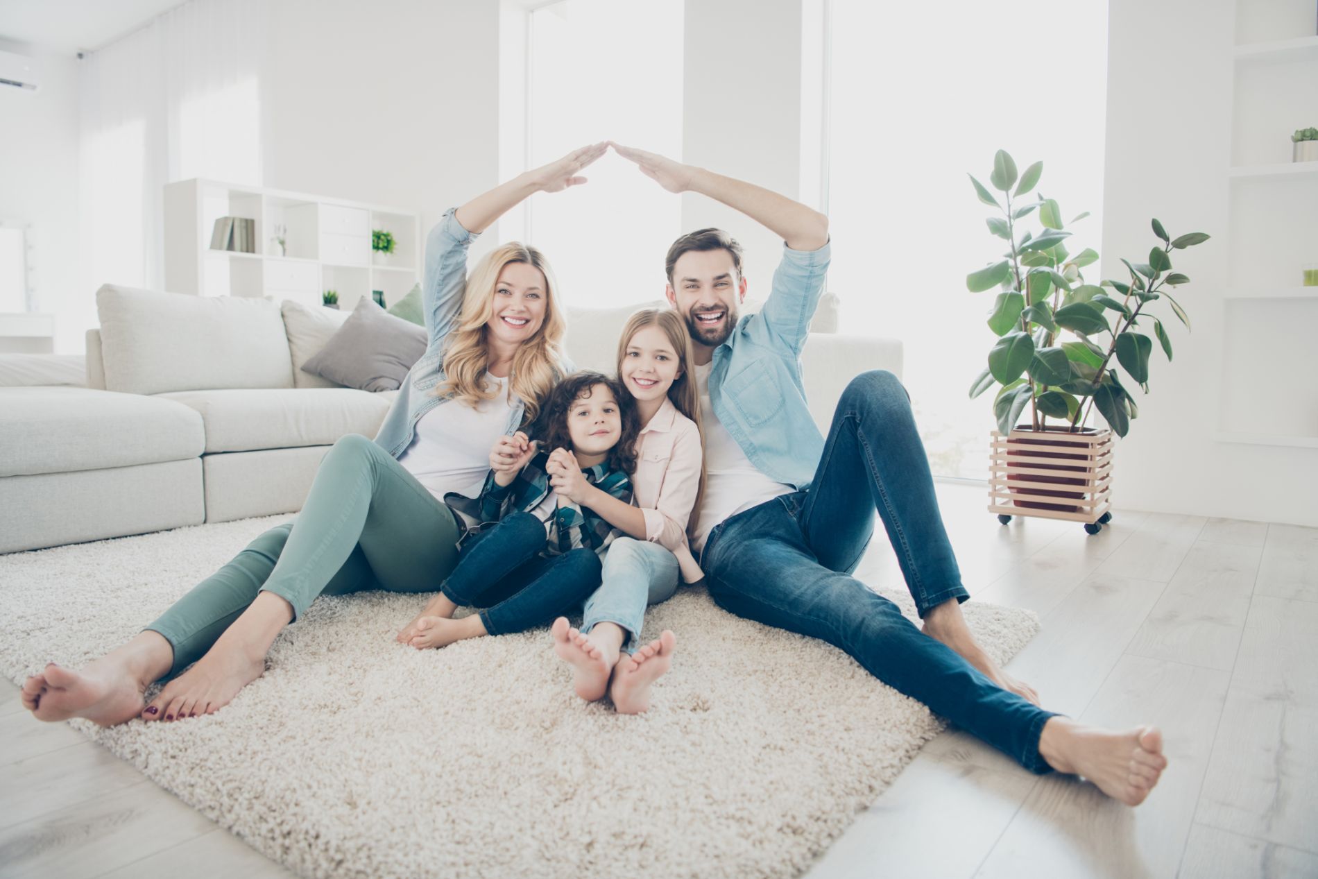 Family of four sitting on a rug, making a roof with their arms, in a bright living room.