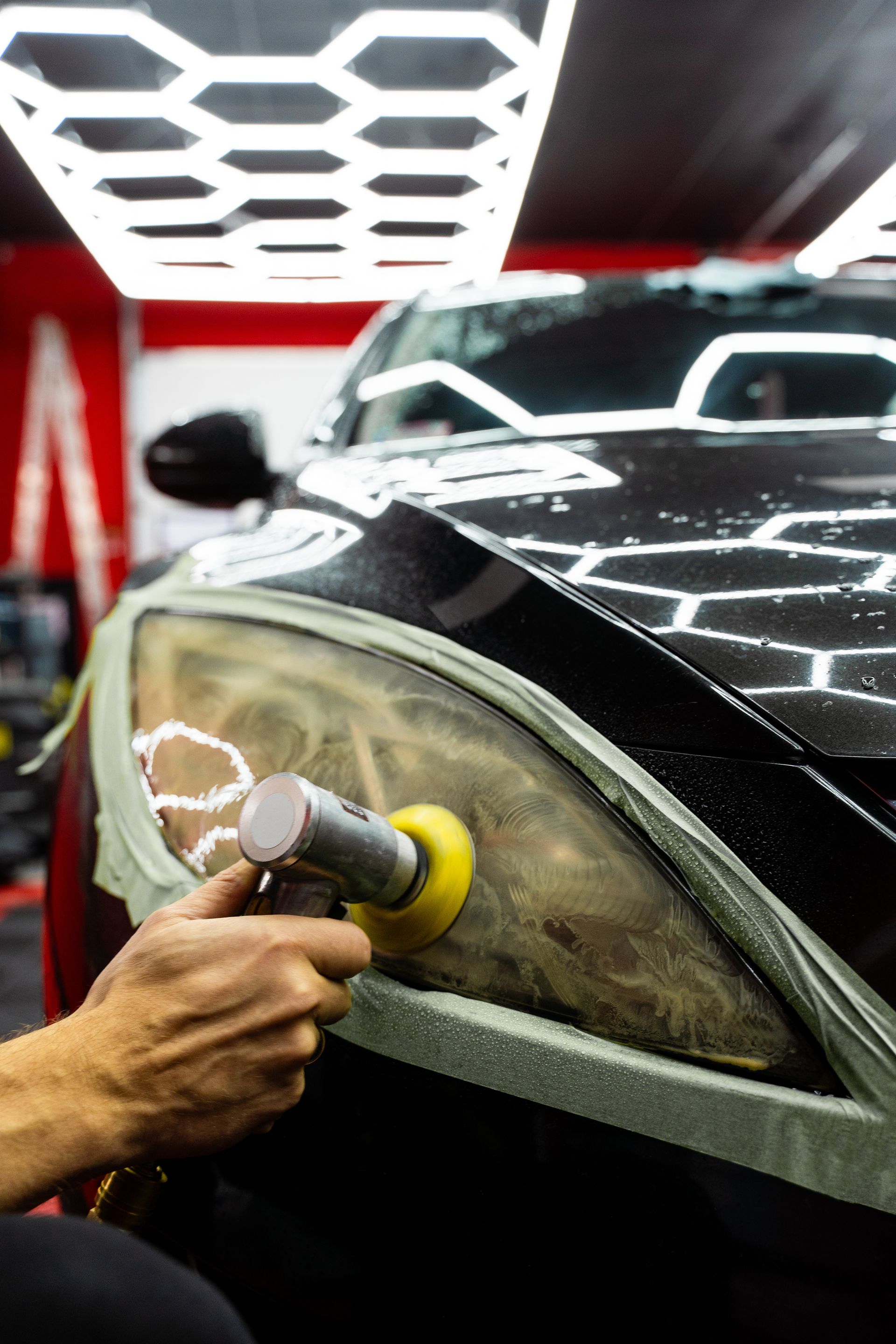 Person polishes a car headlight with a buffing tool. The car is black, the headlight is hazy.