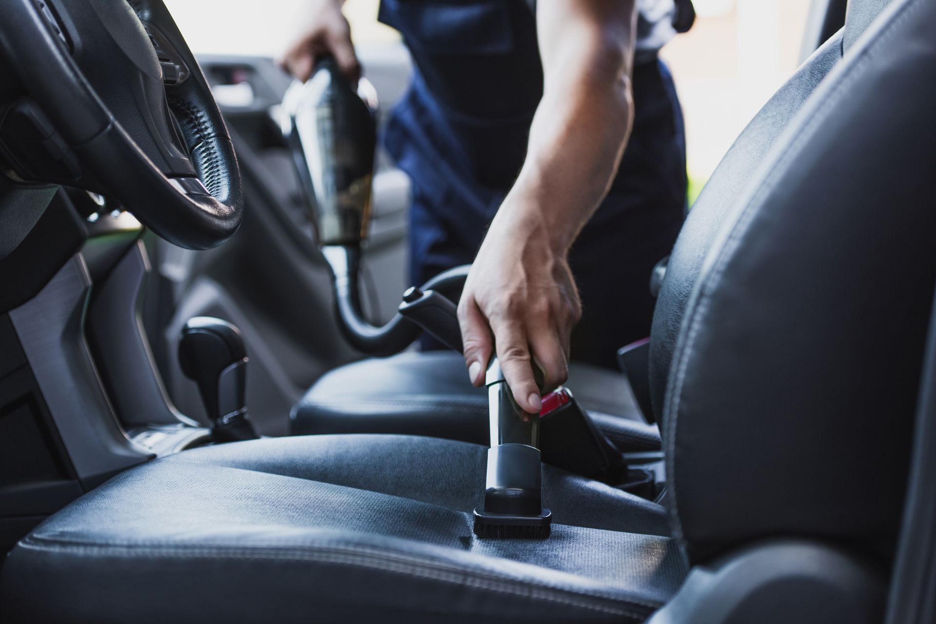 Person vacuuming the interior of a car. Close-up on the black seat, vacuum nozzle pointed down.