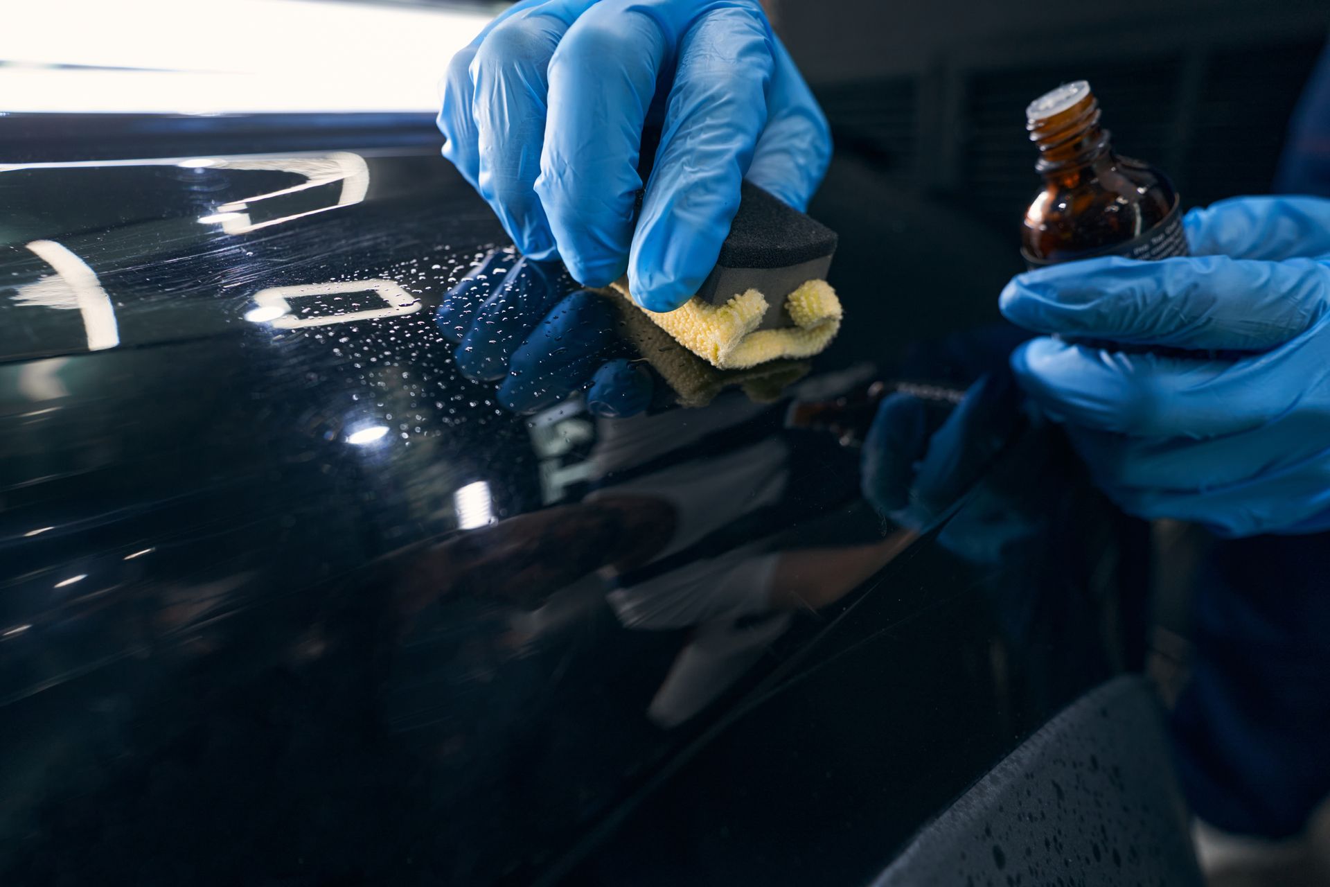 Hands in blue gloves applying car wax to a black car with a sponge.
