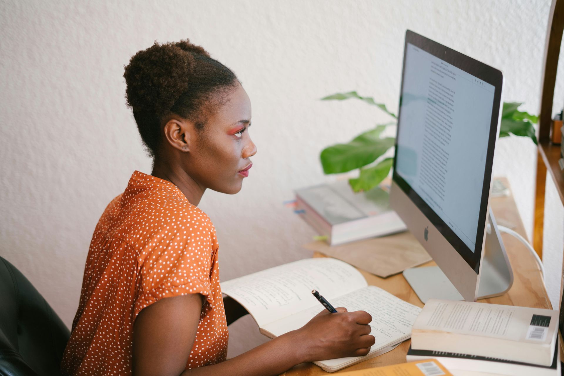 Person writing notes while looking at a desktop computer in a bright office.