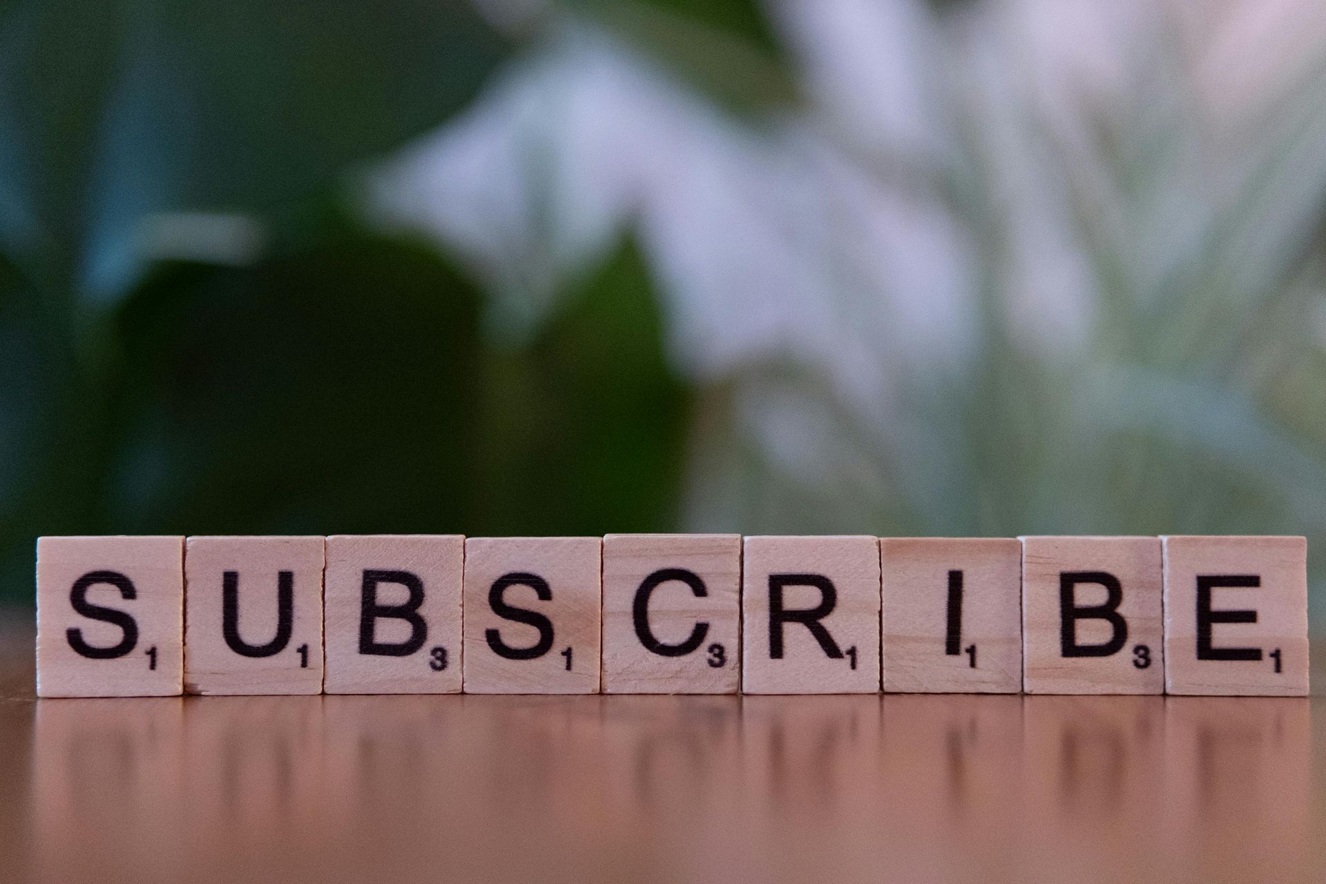 Letter tiles spelling “SUBSCRIBE” on a wooden surface with blurred greenery in the background
