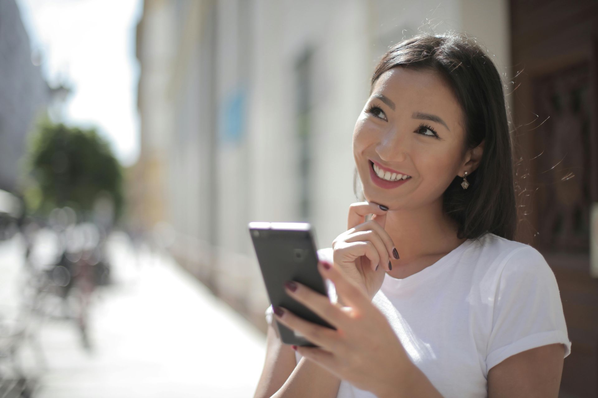 Smiling woman in a white shirt using a smartphone outdoors on a sunny sidewalk
