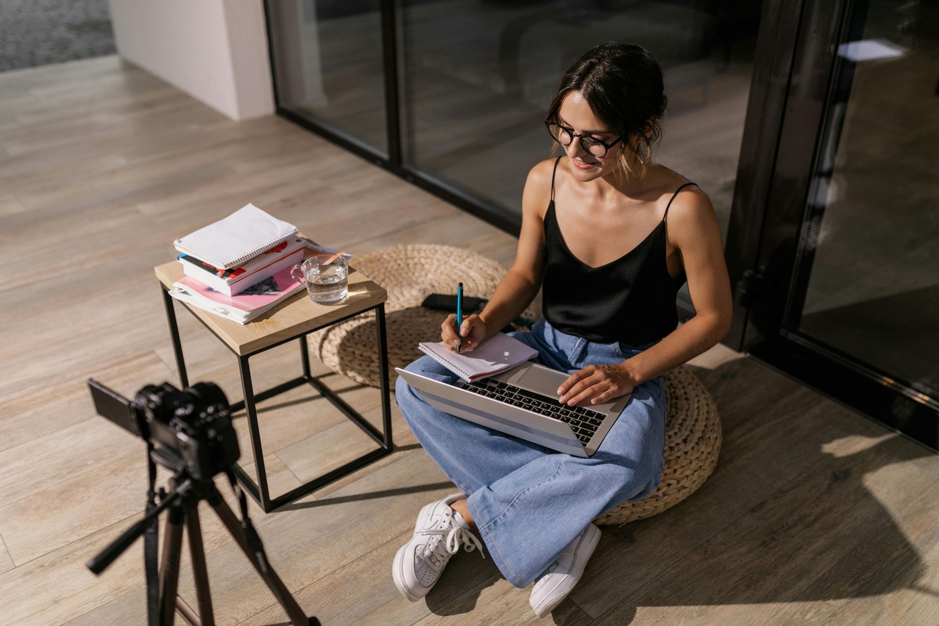 Woman working on a laptop on a floor cushion beside a small table and camera tripod in a bright room