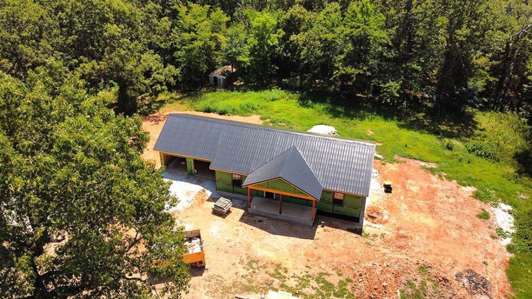 An aerial view of a single-story house under construction with a dark metal roof, set in a clearing surrounded by woods.