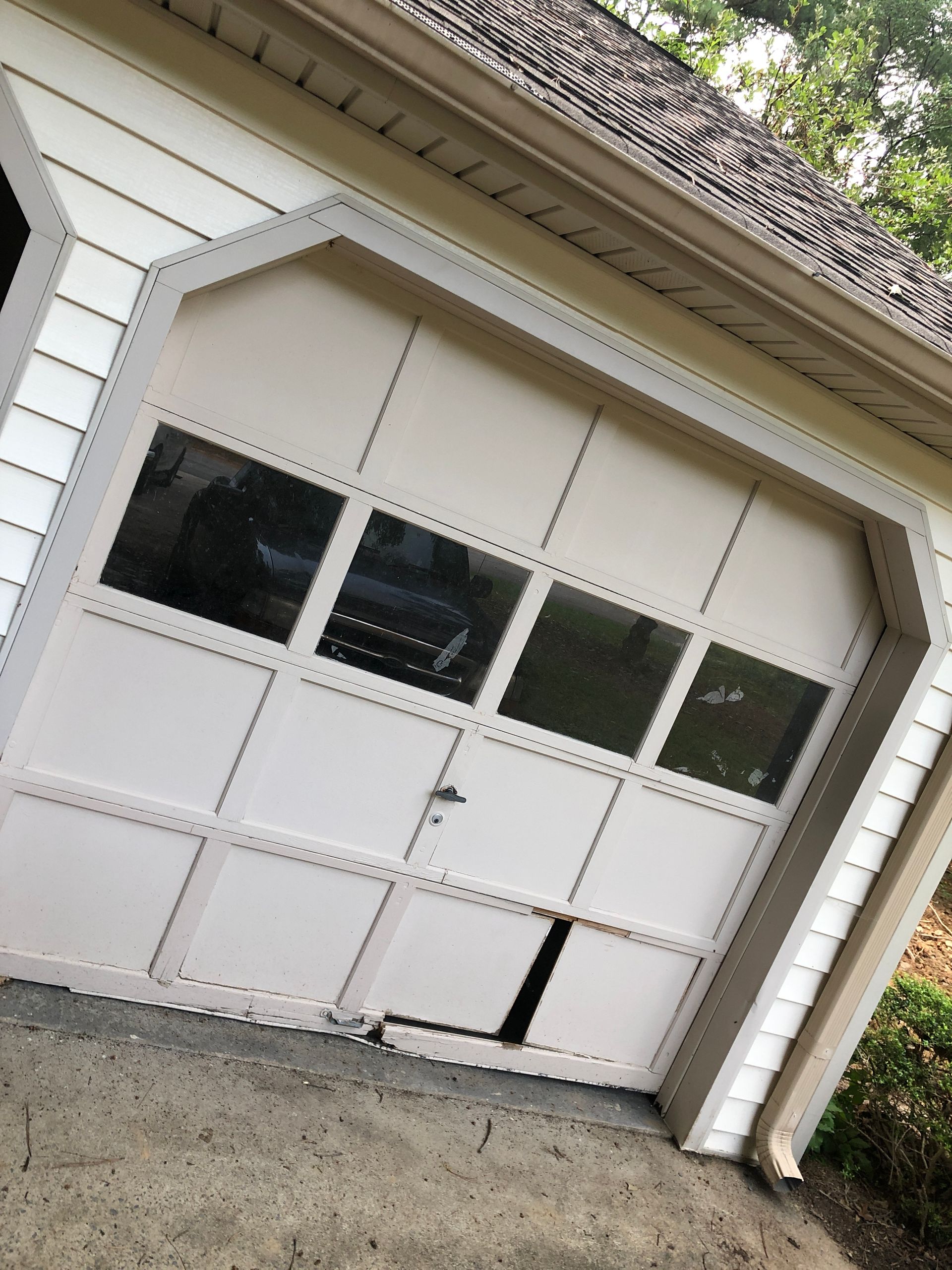 A white, paneled garage door with a row of windows, showing visible damage and a hole in the bottom left section.