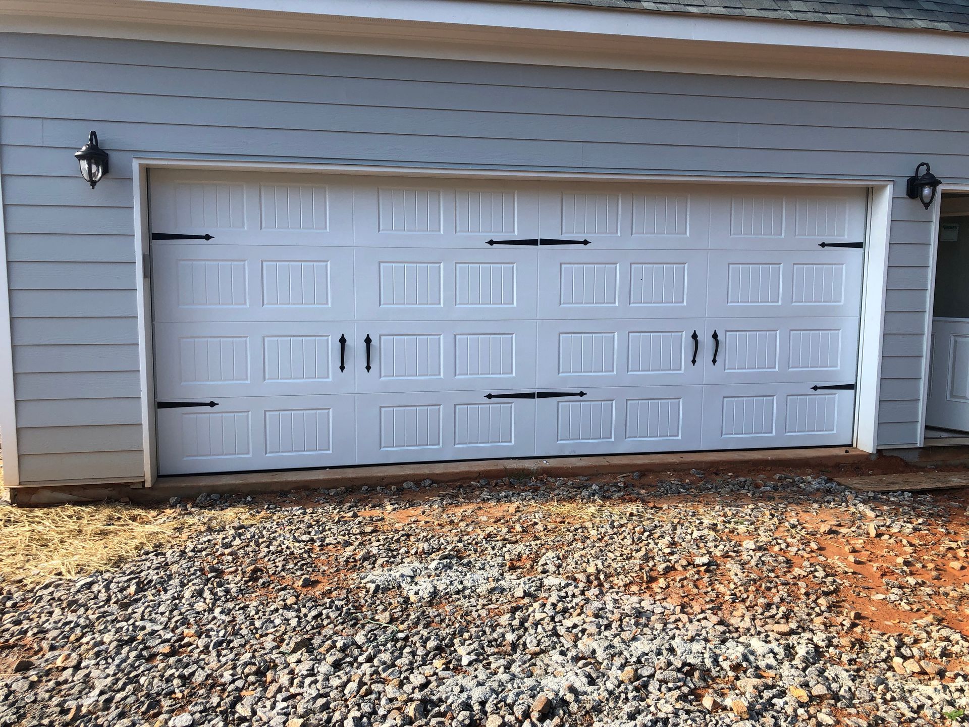 A white, multi-panel garage door with black decorative hinges and handles on a grey-sided house above a gravel driveway.