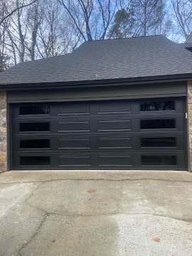 A black garage door with four horizontal windows on each side, set in a stone house exterior with a gray shingle roof.