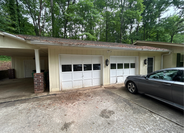 A side-view of a two-car garage with white doors, a covered carport to the left, and a grey car parked in the driveway.