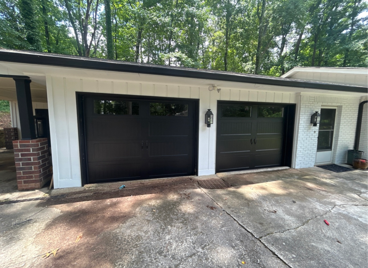 Two black garage doors with windows on a white house exterior with a brick pillar and trees in the background.