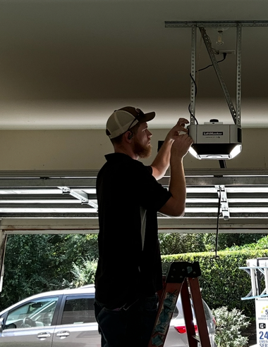 A technician on a ladder repairs a garage door opener mounted to the ceiling.