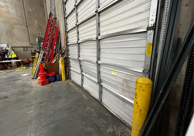 A damaged, dented metal garage door inside a warehouse next to a yellow bollard and stacked ladders.