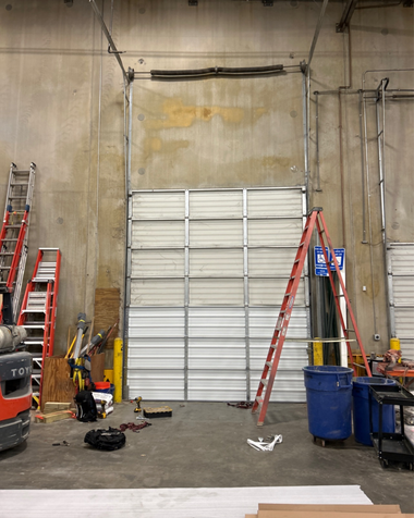 Interior of a warehouse with a partially open industrial garage door, a ladder, and miscellaneous tools on the floor.