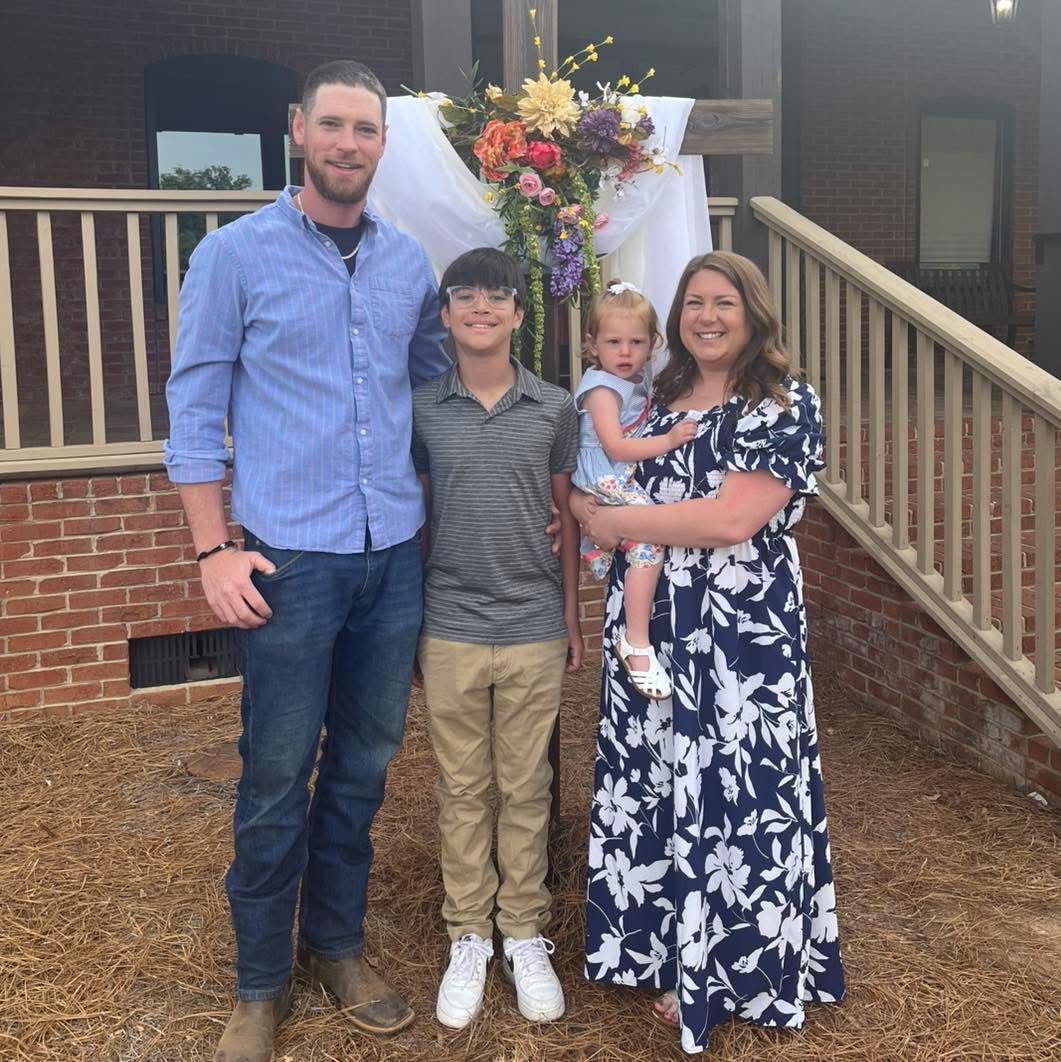 A family of four stands together outside by a decorative cross in front of a brick building.