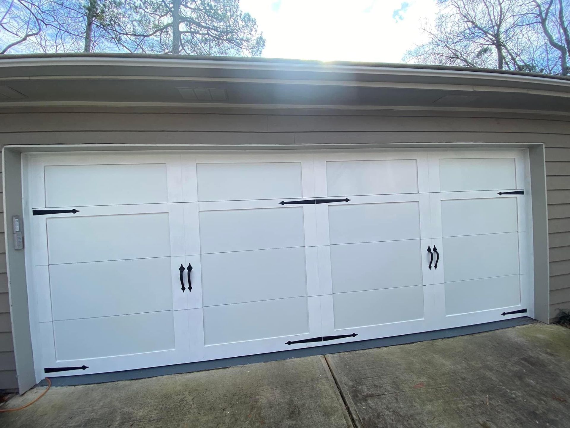 A white four-panel garage door with decorative black iron hinges and handles, installed on a house exterior.