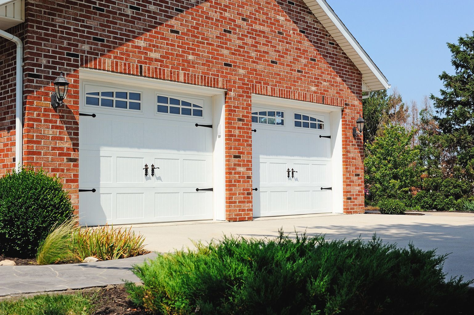 Two white carriage-style garage doors on a brick house exterior with green landscaping in the foreground.